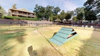 This outdoor amenity space is designed to serve both pets and their owners, offering two distinct but connected areas within a secure, fenced perimeter. The Bark Park is a generously sized, open-air dog park enclosed by sturdy black iron fencing, providing a safe and spacious environment for pets to roam freely. The grounds are covered in grass and feature a green wooden A-frame agility structure, offering enrichment and play opportunities for dogs of all sizes. A bench is positioned along the perimeter fence, giving owners a comfortable place to relax while supervising their pets. Access to the Bark Park is provided through a gated chain-link entry, with concrete steps and a retaining wall guiding visitors from the adjacent pathway down into the space. Mature trees and dense landscaping border the area, providing natural shade and a pleasant, park-like atmosphere. Adjacent to the dog park, the Grill Park area offers outdoor seating with patio chairs and tables, making it an ideal spot for casual gatherings. Signage throughout the space outlines community guidelines for both areas. The surrounding backdrop includes multi-story residential buildings with balconies and a gazebo structure, all framed by a canopy of tall, established trees.
