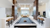 This contemporary amenity space offers an impressive open-plan layout defined by soaring double-height ceilings and an abundance of natural light streaming in through expansive floor-to-ceiling windows at the far end. The design is anchored by a full-size billiards table at the center, finished in a sleek dark felt and framed by tall, wood-paneled columns that add warmth and visual structure to the otherwise bright, neutral interior. Recessed lighting and a statement chandelier overhead provide layered illumination throughout the space. Beyond the billiards area, a comfortable lounge zone features plush cream-toned seating arranged to encourage conversation and relaxation, while a bar-height counter with stools along one side offers a casual perch for socializing. The open sightlines connect each functional zone seamlessly, creating a fluid, cohesive environment. The polished white flooring reflects light throughout, enhancing the sense of spaciousness. Large windows at the rear frame views of the surrounding outdoor environment, further blurring the boundary between interior and exterior. The overall ambiance is refined yet inviting, combining upscale design elements with practical amenities suited for communal gathering and leisure.