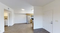 This single-floor residential unit offers a clean, open-concept layout that seamlessly connects the living area to the kitchen. The space is finished with durable wood-look flooring throughout, lending a warm and cohesive feel to the interior. Crisp white walls and flush-mount ceiling lighting keep the atmosphere bright and airy.

The kitchen is a functional highlight of the unit, featuring warm honey-toned wood cabinetry that provides ample storage space. Stainless steel appliances — including a full-size refrigerator, an electric range, and an over-the-range microwave — complete the kitchen's modern and practical setup. A partial wall divides the kitchen from the main living area, creating a sense of definition without sacrificing the open feel of the floor plan.

The living area is generously proportioned, offering flexible space for a variety of furniture arrangements. A white door visible along the wall leads to additional areas of the unit. The overall design is straightforward and well-maintained, making it an accessible and functional space suited for everyday living. The neutral color palette and consistent flooring throughout ensure a unified, move-in-ready aesthetic.