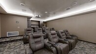 This purpose-built movie room at 28 Seventh St NE in Rochester, Minnesota, delivers a true cinematic experience in a thoughtfully designed, self-contained space. The room is outfitted with rows of plush, dark brown leather power recliner seats, each equipped with built-in cupholders and individual armrests, providing comfortable, theater-style viewing for multiple guests. A ceiling-mounted projector directs onto a large, dedicated projection screen that spans the front wall, creating an immersive big-screen experience. The ceiling features a recessed tray detail accented with continuous LED cove lighting, casting a warm ambient glow throughout the room. Multiple in-ceiling speakers are distributed across the ceiling, supporting a full surround sound audio setup. The walls are finished in warm, deep taupe tones that enhance the darkened atmosphere ideal for screenings. Multi-pattern commercial-grade carpet tiles cover the floor, adding both comfort and acoustic dampening. A built-in dark wood shelving and cabinetry unit along the rear wall provides storage and display space, accompanied by a classic popcorn machine — a charming nod to the traditional movie theater experience. The room is accessed through a solid white paneled door, and smart control panels on the walls allow for convenient management of the room's audiovisual and climate systems.