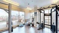 This fitness room is a thoughtfully designed exercise space that combines functionality with refined architectural details. The room features warm hardwood flooring throughout, complemented by crisp white wainscoting that lines the lower walls, crown molding along the ceiling perimeter, and neutral warm-toned walls that create a clean, inviting atmosphere. Flush fluorescent lighting fixtures provide bright, even illumination across the space.

A standout feature of this fitness room is the expansive sliding glass windows along one wall, offering panoramic views of a vibrant urban skyline and access to an outdoor terrace furnished with lounge seating — a rare amenity that allows natural light to fill the room and provides a motivating backdrop during workouts.

The room is well-equipped with a range of fitness equipment, including a Cybex cable machine and weight bench for strength training, a power tower for bodyweight exercises, a BOSU balance trainer, stability balls, and free weights. A wall-mounted flat-screen television adds an entertainment element. A paper towel dispenser near the entry door promotes cleanliness and convenience. The glass-paneled entry door provides visibility into the space. Overall, this fitness room delivers a polished, comfortable, and fully functional environment suited for a comprehensive workout routine.