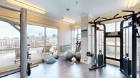 This fitness room is a thoughtfully designed exercise space that combines functionality with refined architectural details. The room features warm hardwood flooring throughout, complemented by crisp white wainscoting that lines the lower walls, crown molding along the ceiling perimeter, and neutral warm-toned walls that create a clean, inviting atmosphere. Flush fluorescent lighting fixtures provide bright, even illumination across the space.

A standout feature of this fitness room is the expansive sliding glass windows along one wall, offering panoramic views of a vibrant urban skyline and access to an outdoor terrace furnished with lounge seating — a rare amenity that allows natural light to fill the room and provides a motivating backdrop during workouts.

The room is well-equipped with a range of fitness equipment, including a Cybex cable machine and weight bench for strength training, a power tower for bodyweight exercises, a BOSU balance trainer, stability balls, and free weights. A wall-mounted flat-screen television adds an entertainment element. A paper towel dispenser near the entry door promotes cleanliness and convenience. The glass-paneled entry door provides visibility into the space. Overall, this fitness room delivers a polished, comfortable, and fully functional environment suited for a comprehensive workout routine.