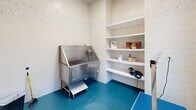 Located at 1931 Chemin Metairie Pkwy in Youngsville, Louisiana, this dedicated pet grooming room is thoughtfully designed to accommodate professional grooming needs within a compact, efficient footprint. The room features white subway tile walls adorned with a decorative mosaic border near the upper portion, lending a clean and polished aesthetic throughout the space. A vibrant teal epoxy floor provides a durable, easy-to-clean surface well-suited for a wet environment. The centerpiece of the room is a stainless steel pet bathing tub equipped with a built-in step for easy pet access, a handheld sprayer, and an enclosed lower cabinet for storage. Adjacent to the tub, a set of built-in white shelves offers organized storage for grooming supplies, products, and tools, including a dryer. A height-adjustable grooming table with a grooming arm provides a dedicated station for finishing work. Natural light filters into the room through a window fitted with horizontal blinds, brightening the otherwise utilitarian space. A standard entry door and wall-mounted fixtures complete the room's functional layout, making this grooming suite a well-equipped and hygienic environment for pet care services.