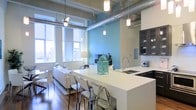 This single-floor space showcases a compelling blend of industrial and contemporary design elements. Exposed silver ductwork runs prominently across the ceiling, complemented by a series of sleek pendant lights that drop at varying heights, creating visual interest overhead. The open-concept layout seamlessly connects the kitchen and living areas, promoting a sense of spaciousness and flow.

The kitchen features a generous white waterfall-edge island with seating for multiple guests, serving as both a functional workspace and a social hub. Dark upper cabinetry with glass fronts provides striking contrast against the light countertops, while a stainless steel sink and modern fixtures complete the culinary space.

The living area is anchored by a plush white sofa and accented by warm hardwood flooring throughout. A vibrant teal accent wall adds a bold pop of color, enhancing the otherwise neutral palette. Large windows along the exterior wall flood the space with abundant natural light while offering views of the surrounding cityscape.

The overall ambiance balances an urban, loft-inspired aesthetic with warm, inviting touches, making the space feel both stylish and comfortable.