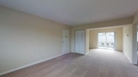 This single-floor space features a open, carpet-floored living area finished in warm, neutral beige tones that create a calm and inviting atmosphere. The main living space is generously proportioned, with smooth white ceilings and clean white baseboards that contrast neatly against the light-colored walls. A set of bifold closet doors offers convenient built-in storage, while a standard entry door provides a clear and functional layout. The space flows naturally toward an adjacent area, likely a dining space, which is illuminated by a elegant chandelier and anchored by a full-width sliding glass door. This glass door floods the area with abundant natural light and opens to a balcony, offering views of the surrounding exterior. The consistent carpeting throughout ties the spaces together cohesively, while the simple, uncluttered design provides a versatile canvas suitable for a variety of furnishing styles. Overall, the space presents a functional and comfortable layout with practical storage solutions, natural light, and seamless indoor-outdoor connectivity through the balcony access.