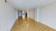 This single-floor space presents an open-concept layout that maximizes the sense of spaciousness throughout the main living area. Light-toned hardwood flooring runs continuously across the entire unit, reflecting overhead lighting and contributing to a bright, airy atmosphere. The textured popcorn ceiling adds a classic finish overhead, while the clean white walls provide a neutral backdrop throughout.

The expansive main living area flows seamlessly toward the kitchen zone, which is partially separated by a low dividing wall that maintains the open feel while defining the cooking space. The kitchen is equipped with stainless steel appliances, including a refrigerator, and features dark countertops that contrast with the lighter surroundings. Overhead pendant lighting illuminates the kitchen area effectively.

A hallway visible from the main living space leads toward additional rooms, with a door visible at the far end suggesting access to a private entrance or utility area. The overall layout is straightforward and functional, with a clean, minimalist aesthetic. The consistent flooring and neutral color palette throughout create a cohesive, well-maintained environment suited to a variety of uses.