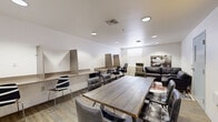 Located at 510 E Third St in Pomona, California, this single-floor multipurpose workspace offers a well-organized and functional layout designed to accommodate both collaborative work and focused individual tasks. The space features wood-look flooring throughout, white walls, and recessed lighting that creates a clean, well-lit environment conducive to productivity.

At the center of the room sits a large rustic wood conference table surrounded by tufted leather chairs, making it ideal for group meetings or collaborative sessions. Along one wall, a continuous built-in desk shelf with divider panels provides individual workstations, each accompanied by ergonomic task chairs — offering dedicated areas for focused, heads-down work.

The lounge area at one end of the space includes a dark leather sofa, a leather armchair with ottoman, and a small round coffee table, all arranged atop a vibrant, multicolored area rug. This seating arrangement provides a relaxed setting for informal discussions or breaks. A compact refrigerator and microwave are also present, adding a practical amenity to the space.

A whiteboard mounted on one wall supports presentations and brainstorming sessions. A decorative illuminated sign adds a touch of character, while a large floral wall print enhances the overall aesthetic. Two entry doors and a window with blinds complete the layout, providing natural light and easy access.