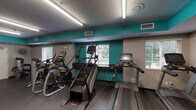 This fitness center is designed to support a well-rounded workout experience within a single-level space. The room is visually defined by bold teal accent walls paired with neutral gray lower walls, creating an energetic yet functional atmosphere. Durable rubber flooring runs throughout, providing a safe and practical surface for all exercise activities.

The cardio area is outfitted with multiple treadmills, elliptical trainers, a stair climber, and a stationary bike, each equipped with individual monitors. Natural light enters through several windows fitted with blinds, offering outdoor views while keeping the space comfortable. Overhead linear fluorescent lighting ensures consistent brightness throughout.

The strength and free-weight area features a full dumbbell rack with a wide range of weights, a flat workout bench, and a cable machine. Floor-to-ceiling mirrors line one wall, enhancing the sense of space and allowing users to monitor their form during exercises. A wall-mounted fan provides additional air circulation, and a flat-screen television is mounted above the mirror wall for entertainment. Keycard access at the entrance and posted facility guidelines reflect a secure, well-maintained environment.