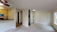 Located at 11733 NE 131st Place in Kirkland, Washington, this single-floor unit offers a well-organized layout designed for comfortable everyday living. The spacious living area is carpeted and filled with natural light, thanks to a large sliding glass door that opens to an outdoor patio area, as well as an additional window. A ceiling fan adds to the comfort of the space, and a built-in shelf provides a subtle architectural detail. Adjacent to the living area, the kitchen features wood-toned cabinetry, dark laminate countertops, a stainless steel double sink, and black appliances including an electric range, over-the-range microwave, and refrigerator. Wood-look flooring grounds the kitchen area. The bedroom area is carpeted and equipped with mirrored sliding closet doors that house a stacked washer and dryer unit, keeping laundry conveniently in-unit. A separate walk-in closet provides generous storage space. The bathroom includes a full tub-shower combination, a wood-toned vanity with a single sink, dark countertop, and a large mirror. Textured ceilings are present throughout the unit, and neutral warm tones on the walls create a cohesive, welcoming ambiance across the entire space.