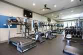 This fitness center is designed to support a full range of workout routines in a well-organized, single-floor layout. The space is finished with durable rubber flooring throughout and features a two-tone wall design with a bold teal accent band contrasting against neutral upper walls, lending the facility a clean and energetic aesthetic. Crown molding and recessed lighting add a polished touch to the interior, while ceiling fans help maintain comfortable airflow.

Large windows along one wall invite natural light and offer views of the surrounding greenery, creating an open and refreshing atmosphere. Glass panel partitions further enhance the sense of space, and floor-to-ceiling mirrors are strategically placed to visually expand the room and assist with form-checking during workouts.

The cardio zone is well-stocked with treadmills, elliptical trainers, and upright and recumbent stationary bikes. A dedicated strength training area houses multiple cable and weight machines, along with a fully stocked free weight rack offering dumbbells and a variety of medicine balls and kettlebells. A flat weight bench provides additional functionality. French doors lead to the exterior, offering convenient access in and out of the facility. A water fountain station is also present, ensuring easy hydration for users.