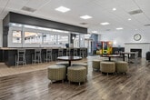 This well-appointed clubhouse offers a generously sized, open-plan layout spread across a single floor, providing ample space for social gatherings and community events. The interior features a contemporary design with a neutral color palette, combining crisp white upper walls with a contrasting dark gray wainscoting that adds depth and sophistication throughout the space.

Flooring transitions seamlessly between light wood-look planks and tile, delineating distinct functional zones. A prominent bar-style counter with bar seating runs along one side, illuminated by pendant lighting and backed by large windows that invite natural light and views of the surrounding building exterior. Adjacent to the counter, vending machines provide added convenience.

The lounge area is thoughtfully furnished with a mix of dark leather sofas, accent chairs, and a wall-mounted flat-screen television, creating a comfortable media and relaxation zone. A fireplace serves as an attractive focal point, adding warmth and character. Dining and gathering areas feature a combination of round tables with patterned ottomans and rectangular tables with bar-height chairs, offering flexible seating arrangements for various group sizes.

The drop ceiling with recessed and flush panel lighting ensures the entire space is evenly and brightly lit. Overall, the clubhouse presents a clean, modern ambiance that is both functional and inviting for residents and guests alike.