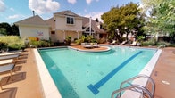 This outdoor swimming pool facility offers a well-appointed recreational retreat designed for relaxation and socializing. The centerpiece is a generously sized, freeform pool with striking blue tile accents and crystal-clear water, complemented by an adjoining circular hot tub. The surrounding deck provides ample space for lounge chairs, with a dedicated row of chaise lounges ideal for sunbathing.

Adjacent to the pool, an elevated wooden deck sits beneath a white pergola supported by classic columns, furnished with outdoor dining and seating arrangements. A separate patio area features a rustic fire pit encircled by cushioned Adirondack chairs, offering a cozy gathering spot with views of the surrounding landscape. Lush greenery, mature trees, and colorful seasonal plantings frame the entire outdoor area, creating a serene, natural backdrop.

The accompanying indoor clubhouse building features herringbone-patterned hardwood flooring, vaulted ceilings with recessed lighting, and an open layout connecting a lounge area with a fireplace and wall-mounted TV to a dining space and a kitchenette with bar seating. Large windows and glass doors allow abundant natural light and direct views of the pool area, seamlessly connecting the indoor and outdoor environments.