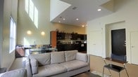 This two-story space showcases a thoughtfully designed open-concept layout that seamlessly integrates the living and kitchen areas into one cohesive environment. The most striking architectural feature is the dramatic double-height ceiling in the main living area, with strategically placed upper-level windows that flood the space with natural light, creating an airy and expansive atmosphere.

The kitchen is well-appointed with dark cabinetry that provides a bold contrast against the light-toned walls. It features open shelving displaying decorative ceramics and glassware, a range, and ample counter space. A kitchen island or bar counter with seating serves as a transitional element between the cooking and living zones, making it ideal for casual dining or entertaining.

The living area centers around a generous light gray sofa, complemented by a textured area rug and accent lighting mounted on the walls. Recessed ceiling lights throughout provide consistent, warm illumination. A doorway visible in the background hints at additional rooms beyond, while the overall color palette of soft greens, whites, and dark wood tones creates a balanced, modern aesthetic throughout the space.