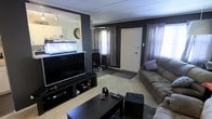 This single-floor home offers a comfortable and characterful living space designed for relaxed everyday living. The main living area makes a strong visual impression with its bold, dark-painted accent walls that contrast with the lighter ceiling and trim, creating a cozy yet defined atmosphere. Soft carpeting underfoot adds warmth throughout the space.

The living room is well-appointed, centered around a large flat-screen television on a media console that provides ample storage for entertainment essentials. Plush, generously sized sofas offer abundant seating, making the room well-suited for both quiet evenings and social gatherings. A sturdy dark coffee table anchors the seating arrangement.

One of the most distinctive features of the home is a built-in aquarium display, seamlessly integrated into the half-wall divider that separates the living area from an adjacent space. This eye-catching element adds life and visual interest to the interior. A wall clock nearby complements the functional character of the room.

Natural light filters into the space through windows dressed with layered curtains, softening the overall ambiance. A front entry door is conveniently accessible from the living area, with a neutral entryway mat marking the transition. Track lighting on the ceiling provides supplemental illumination, ensuring the space remains bright and inviting throughout the day.