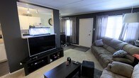 This single-floor home offers a comfortable and characterful living space designed for relaxed everyday living. The main living area makes a strong visual impression with its bold, dark-painted accent walls that contrast with the lighter ceiling and trim, creating a cozy yet defined atmosphere. Soft carpeting underfoot adds warmth throughout the space.

The living room is well-appointed, centered around a large flat-screen television on a media console that provides ample storage for entertainment essentials. Plush, generously sized sofas offer abundant seating, making the room well-suited for both quiet evenings and social gatherings. A sturdy dark coffee table anchors the seating arrangement.

One of the most distinctive features of the home is a built-in aquarium display, seamlessly integrated into the half-wall divider that separates the living area from an adjacent space. This eye-catching element adds life and visual interest to the interior. A wall clock nearby complements the functional character of the room.

Natural light filters into the space through windows dressed with layered curtains, softening the overall ambiance. A front entry door is conveniently accessible from the living area, with a neutral entryway mat marking the transition. Track lighting on the ceiling provides supplemental illumination, ensuring the space remains bright and inviting throughout the day.