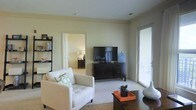This single-floor residence offers a welcoming and well-appointed interior designed for comfortable everyday living. The living room serves as the heart of the space, featuring soft, neutral wall tones and plush carpeting that create a warm and inviting ambiance. A generously sized white sofa anchors the seating area, complemented by a dark wood media console that grounds the room with a touch of contrast. A leaning bookshelf along the wall provides both storage and display space, adding a layer of personality to the room. Natural light filters in through a glazed patio door fitted with blinds, which opens to an outdoor area and enhances the sense of openness throughout the living space. Crown molding along the ceiling adds a refined architectural detail that elevates the overall aesthetic. An open doorway connects the living room to an adjoining bedroom, which is visible in the background and features its own furnishings, maintaining a cohesive and calm design palette throughout. The overall ambiance is clean, understated, and functional, making it a comfortable and practical living environment.