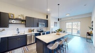 This thoughtfully designed 1-bedroom, 1-bathroom residence offers a cohesive open-concept layout that maximizes every square foot across a single floor. The kitchen serves as the centerpiece of the living space, showcasing bold navy cabinetry, a textured white hexagon tile backsplash, a full suite of stainless steel appliances, and a large island with a quartz countertop and bar seating — ideal for both meal preparation and casual dining. Pendant lighting above the island adds a modern touch, while light wood-style flooring flows seamlessly throughout the main living area. The living room is bright and airy, with access to a private balcony that brings in ample natural light and outdoor views. The bedroom is a comfortable, carpeted retreat featuring a large window that further enhances the sense of light and openness. The full bathroom is finished with wood-look tile flooring, dark cabinetry with gold hardware, a white vanity countertop, and a tub-shower combination. A dedicated in-unit laundry area houses a full-size top-loading washer and dryer with overhead wire shelving for added storage. Throughout the space, neutral warm tones and clean lines create a modern, inviting ambiance.