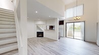 This two-story home offers a well-organized layout across two floors, with the majority of the living space situated on the main level. The open-concept ground floor seamlessly connects the living room and kitchen, creating an airy, functional space bathed in natural light from large sliding glass doors that lead to a private balcony with outdoor views. A standout feature of the living area is the wood-burning fireplace, framed by a clean white surround and nestled beneath the staircase, adding both character and warmth to the space. Soaring double-height ceilings above the living room enhance the sense of openness. Light wood-tone laminate flooring runs consistently throughout both floors, lending a cohesive and modern aesthetic. The galley-style kitchen is efficiently designed, featuring rich dark wood cabinetry, granite-look countertops, a stainless steel sink, and full-size appliances. A half bathroom on the main floor provides added convenience. The staircase, finished in crisp white with open railings, leads to the upper level, where the spacious primary bedroom offers recessed lighting, a dedicated closet, and its own full bathroom complete with a walk-in shower, white vanity, and mirror. A second full bathroom on this floor features a granite-top vanity and natural light from a frosted window.