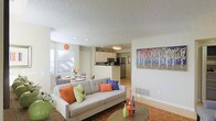 This single-floor residence offers a well-designed open-concept layout that seamlessly connects the living, dining, and kitchen areas into one cohesive and functional space. The living area is anchored by a comfortable sofa adorned with vibrant accent pillows in orange, blue, and green, complemented by a patterned area rug that adds warmth and personality to the room. A textured popcorn ceiling with recessed lighting runs throughout, providing a bright and airy atmosphere. Large windows with blinds invite ample natural light, enhancing the neutral beige and white color palette of the walls. The dining area sits adjacent to the living space, offering an intimate setting ideal for everyday meals or entertaining. The kitchen features white cabinetry, dark countertops, and stainless steel appliances including a refrigerator and range, all centered around a convenient breakfast bar that doubles as a transitional element between the kitchen and living areas. A colorful piece of wall art depicting a vibrant tree scene adds a striking focal point to the main living area, tying together the home's lively yet balanced aesthetic. The overall ambiance is warm, inviting, and thoughtfully arranged for comfortable everyday living.