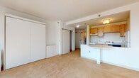This single-floor unit is organized around an open-concept layout that connects the living area to the kitchen, creating a functional and cohesive living space. The living area is finished with wall-to-wall beige carpeting and features a large built-in wardrobe along one wall, accompanied by a compact dresser. A sliding closet door provides additional storage access, and the textured ceiling adds subtle visual depth throughout the space.

The kitchen is positioned along one side of the open floor plan and is defined by a curved, white peninsula that doubles as a breakfast bar. Warm-toned wooden cabinetry lines the walls, complemented by a white electric range, a dishwasher, and a full-sized refrigerator. A range hood is mounted above the stove, and the kitchen flows seamlessly into the main living space.

The bedroom is a generously sized, light-filled room with carpeted floors and a large window that frames views of a nearby urban streetscape, allowing natural light to fill the room throughout the day.

The bathroom features stone-look tile flooring and includes a full soaking tub with an overhead shower. A washer unit is conveniently integrated into the bathroom, along with a vanity with light wood cabinetry and a white countertop.
