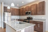 Located at 7920 Watts Rd in Madison, WI, this one-bedroom, two-bathroom unit offers a well-designed single-floor layout that maximizes every square foot. The kitchen is a standout, featuring rich dark-toned cabinetry extending to the ceiling for ample storage, granite countertops, and a full suite of stainless steel appliances including a refrigerator, range, and built-in microwave. A central island with a granite surface and integrated sink provides additional prep space and casual seating, illuminated by dual pendant lights overhead. Light wood-style flooring runs throughout the kitchen and living areas, creating a warm, cohesive aesthetic. The living room opens directly to a sliding glass door fitted with vertical blinds, flooding the space with natural light and providing views of the surrounding greenery. The bedroom is carpeted for comfort and features a ceiling fan, a large window with outdoor views, and a dedicated closet. Two full bathrooms each include a tub/shower combo and are finished with tile flooring and vanity storage. A connecting hallway ties the layout together efficiently, making this a functional and well-appointed residential space.