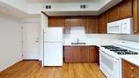 This single-floor residence offers a practical and comfortable layout designed with everyday living in mind. The kitchen is a standout feature, showcasing rich medium-toned wood cabinetry that extends to the upper walls, providing generous storage throughout. White appliances — including a gas range with overhead microwave, refrigerator, and double-basin sink — are set against light countertops, creating a clean, functional workspace. Light wood-tone flooring runs throughout the kitchen and living areas, adding warmth to the neutral palette.

The open living area adjoins the kitchen, offering a generous, flexible space with crisp white walls and the same wood-tone flooring. Natural light enters through a window near the entry door, which also provides views of the surrounding neighborhood. A doorway from the living area leads to a carpeted bedroom, offering a softer underfoot experience suited for rest.

The bathroom features light tile flooring, a vanity with wood cabinetry and a white countertop, and a notable accessibility-oriented grab bar mounted along the wall. Overhead ventilation and clean white walls complete the space. Overall, the unit presents a straightforward, well-maintained interior with consistent finishes throughout.