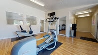 This single-story fitness center is thoughtfully designed to support a well-rounded workout experience. The space is organized across two connected areas, allowing for clear separation between cardio and strength training zones. Warm hardwood flooring runs throughout both sections, complementing the bright white walls and creating a clean, energetic atmosphere.

The main cardio area is outfitted with Precor elliptical trainers, a recumbent bike with an integrated display, and a treadmill, each positioned on protective rubber mats. Fluorescent overhead lighting ensures consistent illumination throughout the space. A wall-mounted flat-screen television adds entertainment options for users during workouts.

A doorway connects to a secondary room that houses a multi-station cable weight machine, providing options for a variety of strength training exercises. A large framed mirror in this area enhances the sense of space and allows users to monitor their form. An exit door offers convenient access to adjacent areas.

Natural light filters in through slatted windows in the main room, while a floor-to-ceiling glass sliding door in a separate nook opens to views of a bright outdoor pool and lounge area. A comfortable armchair positioned near this window offers a relaxing vantage point. The overall ambiance is functional, well-lit, and welcoming.