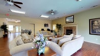 This single-floor space offers a comfortable and inviting atmosphere centered around a generously sized living area. The room is anchored by a classic stone fireplace framed with a clean white mantel, serving as a natural focal point. Recessed lighting and skylights work in tandem to fill the space with bright, even illumination, while a ceiling fan adds both function and a casual charm overhead. The warm hardwood flooring adds richness and continuity throughout. Ample seating is arranged thoughtfully around a wooden coffee table, creating an ideal setting for relaxation or conversation. Decorative touches — including a vase of fresh flowers, indoor potted plants, and framed wall art — add life and personality to the room. A built-in bookshelf near the entry door provides practical storage and hints at a lived-in, curated character. Soft, neutral tones across the walls, furniture, and décor lend the space a cohesive, airy feel. A mirror above the fireplace further enhances the sense of openness. An adjacent area, visible through an open passageway, suggests additional functional space, making this a well-connected and versatile single-floor layout.