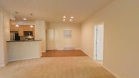 This single-floor space offers a welcoming and functional layout designed around an open-concept living area. The main living space features a combination of warm hardwood flooring and plush carpet, creating distinct zones while maintaining a cohesive and comfortable feel throughout. Recessed lighting in the ceiling provides even, ambient illumination that enhances the bright and airy atmosphere of the room. The walls are finished in a soft, neutral tone, lending a clean and versatile backdrop suited to a variety of interior styles. A baseboard heater along the far wall ensures efficient climate control. Natural light filters in through a window fitted with blinds, adding to the openness of the space. The kitchen is situated to one side and features overhead cabinetry, under-cabinet lighting, built-in appliances including a microwave, and a raised breakfast bar that visually separates it from the living area while maintaining an open connection. Multiple interior doors provide access to additional rooms, offering a practical and well-organized flow throughout the space. White trim and door frames throughout add a crisp, polished finishing touch. Overall, this space combines everyday functionality with a light, inviting ambiance.