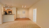 This single-floor space offers a welcoming and functional layout designed around an open-concept living area. The main living space features a combination of warm hardwood flooring and plush carpet, creating distinct zones while maintaining a cohesive and comfortable feel throughout. Recessed lighting in the ceiling provides even, ambient illumination that enhances the bright and airy atmosphere of the room. The walls are finished in a soft, neutral tone, lending a clean and versatile backdrop suited to a variety of interior styles. A baseboard heater along the far wall ensures efficient climate control. Natural light filters in through a window fitted with blinds, adding to the openness of the space. The kitchen is situated to one side and features overhead cabinetry, under-cabinet lighting, built-in appliances including a microwave, and a raised breakfast bar that visually separates it from the living area while maintaining an open connection. Multiple interior doors provide access to additional rooms, offering a practical and well-organized flow throughout the space. White trim and door frames throughout add a crisp, polished finishing touch. Overall, this space combines everyday functionality with a light, inviting ambiance.