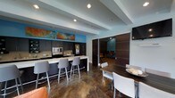 This single-floor space offers a thoughtfully designed communal environment that balances functionality with modern aesthetics. The open-concept layout connects a fully equipped kitchenette to a comfortable dining and lounge area, creating a seamless flow throughout the space.

The kitchenette is outfitted with dark cabinetry, a stainless steel microwave, a full-size refrigerator, and a light-toned stone countertop. A breakfast bar extends from the counter, accompanied by a row of contemporary gray upholstered barstools, providing casual seating for multiple guests. Vibrant abstract artwork adorns the wall above the cabinetry, adding a bold pop of color against the soft blue accent walls.

The dining area features a dark wood table paired with modern upholstered chairs, creating an inviting space for shared meals or collaborative gatherings. The polished concrete flooring throughout lends an industrial yet refined character to the space.

A set of large sliding dark wood panel doors leads to an adjoining lounge room, visible in the background, which offers additional seating in a more intimate setting. Recessed lighting is installed throughout, ensuring even, well-distributed illumination across all areas. The combination of cool blue tones, warm wood accents, and polished surfaces gives this space a clean, contemporary ambiance.