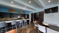 This single-floor space offers a thoughtfully designed communal environment that balances functionality with modern aesthetics. The open-concept layout connects a fully equipped kitchenette to a comfortable dining and lounge area, creating a seamless flow throughout the space.

The kitchenette is outfitted with dark cabinetry, a stainless steel microwave, a full-size refrigerator, and a light-toned stone countertop. A breakfast bar extends from the counter, accompanied by a row of contemporary gray upholstered barstools, providing casual seating for multiple guests. Vibrant abstract artwork adorns the wall above the cabinetry, adding a bold pop of color against the soft blue accent walls.

The dining area features a dark wood table paired with modern upholstered chairs, creating an inviting space for shared meals or collaborative gatherings. The polished concrete flooring throughout lends an industrial yet refined character to the space.

A set of large sliding dark wood panel doors leads to an adjoining lounge room, visible in the background, which offers additional seating in a more intimate setting. Recessed lighting is installed throughout, ensuring even, well-distributed illumination across all areas. The combination of cool blue tones, warm wood accents, and polished surfaces gives this space a clean, contemporary ambiance.