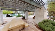 This inviting outdoor courtyard space is defined by its prominent pergola structure, supported by sturdy concrete masonry block columns that frame the patio area with a strong architectural presence. The slatted overhead pergola provides partial shade while allowing natural light to filter through, creating a comfortable environment for outdoor gatherings. Beneath the pergola, large stone benches and a picnic table offer ample seating, making it well-suited for communal use. A vending machine is conveniently positioned along one edge of the covered area. The surrounding landscape is thoughtfully designed, featuring ornamental shrubs, small deciduous trees with seasonal foliage, and raised planting beds bordered by masonry walls. An open-air dining area with a round metal table and chairs is nestled under a mature shade tree, providing a relaxed setting. The broader complex is characterized by white-painted exterior walls, cedar shake roofing accents, and green metal railings on upper-level walkways, all contributing to a cohesive mid-century aesthetic. Concrete pathways connect the various zones of the courtyard, tying together the landscaped beds, seating areas, and building entrances into a unified, functional outdoor environment.