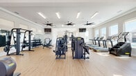 Located in Celina, TX, this Yardly fitness center offers a generously sized, single-floor exercise space designed to support a full range of workout routines. The interior features light hardwood-style flooring throughout, crisp white walls, and tray ceilings that lend a refined, polished aesthetic to the space. Multiple large-blade ceiling fans are suspended overhead alongside linear LED light fixtures, ensuring excellent airflow and consistent illumination across the entire floor.

Natural light pours in through numerous windows lining the perimeter, providing views of the surrounding neighborhood and creating an open, energizing atmosphere. Glass-paneled entry doors at one end further enhance the connection to the outdoors.

The facility is comprehensively equipped with a wide array of commercial-grade Technogym machines, including cable stations, chest and leg press units, and multi-use strength systems. A dedicated free weights area features a full dumbbell rack and adjustable benches. The cardio section is stocked with treadmills, ellipticals, stationary bikes, and a rowing machine. A functional training zone includes medicine balls, kettlebells, TRX straps, and an open floor area ideal for stretching or group exercise. Wall-mounted mirrors and TVs, a water fountain, and the Yardly-branded wall art complete the well-appointed interior.