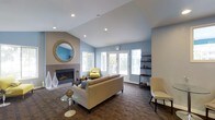 This single-story space centers around a well-appointed living area designed with both comfort and style in mind. The room immediately draws attention to its vaulted ceiling, which adds a sense of spaciousness and volume overhead. Recessed lighting is integrated throughout the ceiling, complementing the generous natural light that streams in through multiple windows and a set of French doors along the far wall. These doors offer a seamless visual connection to the outdoors while brightening the interior with a soft, airy glow.

The living area features a tiled fireplace framed by a clean, modern surround, with a decorative round mirror mounted above it, adding depth and visual interest to the wall. The flooring consists of rich, dark patterned tile that provides a grounded contrast to the soft blue and warm taupe tones of the walls.

Furnishings include a neutral-toned sofa, accent chairs in muted yellow and cream, and a glass-topped side table, creating a relaxed yet polished seating arrangement. A dark wall-mounted shelving unit on the opposite side of the room provides functional display space. The overall ambiance is calm, modern, and inviting.
