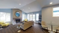 This single-story space centers around a well-appointed living area designed with both comfort and style in mind. The room immediately draws attention to its vaulted ceiling, which adds a sense of spaciousness and volume overhead. Recessed lighting is integrated throughout the ceiling, complementing the generous natural light that streams in through multiple windows and a set of French doors along the far wall. These doors offer a seamless visual connection to the outdoors while brightening the interior with a soft, airy glow.

The living area features a tiled fireplace framed by a clean, modern surround, with a decorative round mirror mounted above it, adding depth and visual interest to the wall. The flooring consists of rich, dark patterned tile that provides a grounded contrast to the soft blue and warm taupe tones of the walls.

Furnishings include a neutral-toned sofa, accent chairs in muted yellow and cream, and a glass-topped side table, creating a relaxed yet polished seating arrangement. A dark wall-mounted shelving unit on the opposite side of the room provides functional display space. The overall ambiance is calm, modern, and inviting.