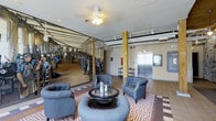 This single-floor space offers a compelling fusion of historical character and contemporary design, making for a memorable and inviting environment. The lobby's most striking feature is a large-scale black-and-white mural depicting a historic industrial scene, likely referencing the building's mill or factory origins, which spans an entire wall and immediately draws the eye upon entry.

Exposed wooden columns punctuate the open floor plan, reinforcing the industrial heritage of the space while adding warmth and architectural interest. The ceiling retains a utilitarian aesthetic with visible track lighting and industrial-style fixtures, complemented by a ceiling fan that adds a residential touch.

The seating area is thoughtfully arranged, featuring tufted gray club chairs and a round glass-topped coffee table, all anchored by a bold geometric-patterned area rug in red and white tones. This creates a comfortable and visually dynamic gathering zone within the larger open space.

The flooring transitions from classic terracotta tile to the patterned rug, adding texture and visual contrast. A stainless steel elevator is visible in the background, underscoring the space's functional, multi-use nature. Overall, the space exudes an atmosphere that honors its industrial past while providing a welcoming, well-appointed environment for its occupants.
