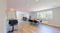 This well-appointed single-story space offers a thoughtfully designed layout encompassing an open-plan kitchen and living area, a separate living room, and a full bathroom. The main gathering area is bright and spacious, featuring light hardwood-style flooring that runs throughout the entire space, recessed ceiling lighting, and multiple windows that invite generous amounts of natural light and offer views of the surrounding greenery outdoors. The open area is configured to support both collaborative work and casual gatherings, with multiple tables and seating arrangements that make it highly functional and adaptable. A ceiling fan adds comfort to the main area. The kitchen is neatly designed with white cabinetry, granite-look countertops, a gas range, a full-size refrigerator, and a sink — all accessible via a pass-through counter that connects seamlessly to the main living area. The separate living room provides a more intimate setting, furnished with a sofa, an armchair, a wall-mounted television, and a ceiling fan, alongside windows that bring in ample natural light. The bathroom is clean and functional, featuring a vanity with white cabinetry, a toilet, and a bathtub with shower. Together, these spaces create a comfortable, multi-use environment with a clean, modern aesthetic.