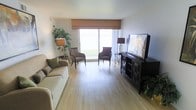 This single-floor living space offers a welcoming and well-organized layout centered around a luminous main living area. Light hardwood flooring runs throughout, lending warmth and a sense of continuity to the space. A large sliding glass door dominates the far wall, flooding the room with natural light and providing an open view to the outdoors, creating an airy, expansive feel. A neutral-toned Roman shade offers privacy while complementing the room's understated color palette.

The living area is thoughtfully arranged with a plush sofa positioned along one wall, paired with two accent chairs near the glass door — an ideal setup for both relaxation and conversation. A dark wood media console anchors the opposite wall, housing a large flat-screen television and providing ample storage. Decorative touches, including floor lamps, a potted plant, and tasteful artwork, add character without overwhelming the space.

The overall ambiance is calm and neutral, with a balance of natural and artificial lighting. The clean lines, light tones, and uncluttered arrangement contribute to a functional yet comfortable living environment.