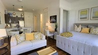 This single-floor space showcases a thoughtfully designed open-concept layout that seamlessly connects the living area to the kitchen. The living room serves as the focal point, featuring rich dark hardwood flooring that contrasts elegantly with the neutral white and light gray walls. Two plush sofas anchored by yellow accent pillows create a welcoming seating arrangement, complemented by a tray coffee table and decorative wall art. A compact writing desk and chair occupy a functional nook along one wall, making efficient use of the available space. Pendant lighting and a table lamp provide layered illumination throughout. The kitchen, visible from the living area, features dark cabinetry and is accompanied by a glass-top dining table. White interior doors lead to additional areas, including what appears to be a hallway and closet spaces. The overall aesthetic is contemporary and cohesive, with a yellow and gray color palette lending warmth and personality to the clean, modern design. The efficient floor plan maximizes every corner while maintaining an open and airy feel throughout.