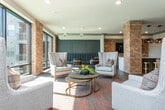 Located in Atlanta's Bolton Drive corridor, this thoughtfully designed single-floor community amenity space blends industrial warmth with modern sophistication. Exposed brick columns serve as defining architectural anchors throughout, providing a rich textural contrast against smooth, neutral-toned walls and wood-look tile flooring that flows seamlessly across the entire space.

At the heart of the room, a generously sized island with a dramatic veined marble countertop commands attention, surrounded by sleek black-and-gold barstools beneath a trio of woven pendant lights. The wet bar area behind the island features gray shaker cabinetry, a wall-mounted television, and subway tile backsplash — ideal for social gatherings or casual entertaining.

The lounge area transitions naturally from the bar, offering an arrangement of elegant wingback chairs and a round coffee table centered on a richly patterned area rug. Floor-to-ceiling black-framed windows and glass doors flood this zone with natural light while providing unobstructed views of the outdoor pool and surrounding community.

A separate dining nook with mid-century modern chairs and round tables adds a functional layer to the space. Curated artwork adorns the walls, and recessed lighting throughout ensures a warm, inviting ambiance at any hour. The overall design achieves a refined yet approachable atmosphere suited for both relaxation and community interaction.