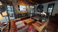 Located at 12103 Inwood Cir in Dallas, Texas, this single-story lounge space presents a carefully curated blend of industrial and upscale design elements. The architectural framework is defined by prominent exposed wood beam ceilings, rich hardwood flooring, and expansive floor-to-ceiling steel-framed windows that flood the interior with abundant natural light while offering views of the surrounding landscaping.

A dramatic geometric wood-paneled accent wall serves as the room's focal point, anchoring a sleek linear fireplace and a mounted display screen above it. The seating area is anchored by classic tufted Chesterfield sofas and leather wingback chairs arranged around a gold-accented coffee table atop a vibrant Persian-style rug, creating a warm and inviting atmosphere.

An industrial-style chandelier with exposed Edison bulbs hangs above the central seating arrangement, adding to the space's layered lighting design. Steel-framed glass partition doors with black mullions lead to an adjacent lounge area with patterned tile flooring, a bar counter, and pendant lighting, creating a distinct yet connected secondary gathering space. An arched interior window further adds architectural character, contributing to the overall sophisticated and welcoming ambiance throughout.