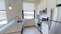 This single-floor, one-bedroom, one-bathroom residence features a thoughtfully designed kitchen as a central highlight. The kitchen is outfitted with white shaker-style cabinetry that extends to the upper walls, providing ample storage throughout. Granite countertops with a speckled grey tone add a polished, durable work surface, complemented by a stainless steel undermount sink with a modern arc faucet. The kitchen is equipped with a full stainless steel appliance package, including an electric range with oven, an over-the-range microwave, and a dishwasher. Light wood-tone flooring runs throughout, lending warmth to the space. A large window with a roller shade allows natural light to fill the kitchen while framing an urban rooftop view of the surrounding cityscape. The clean white walls and neutral finishes create a bright, functional atmosphere. The efficient galley-style layout maximizes the available space, making it practical for everyday living. Overall, the unit presents a clean, contemporary aesthetic suited for urban residential living.