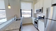This single-floor, one-bedroom, one-bathroom residence features a thoughtfully designed kitchen as a central highlight. The kitchen is outfitted with white shaker-style cabinetry that extends to the upper walls, providing ample storage throughout. Granite countertops with a speckled grey tone add a polished, durable work surface, complemented by a stainless steel undermount sink with a modern arc faucet. The kitchen is equipped with a full stainless steel appliance package, including an electric range with oven, an over-the-range microwave, and a dishwasher. Light wood-tone flooring runs throughout, lending warmth to the space. A large window with a roller shade allows natural light to fill the kitchen while framing an urban rooftop view of the surrounding cityscape. The clean white walls and neutral finishes create a bright, functional atmosphere. The efficient galley-style layout maximizes the available space, making it practical for everyday living. Overall, the unit presents a clean, contemporary aesthetic suited for urban residential living.
