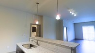 This single-floor space showcases a thoughtfully designed open-concept layout that seamlessly connects the kitchen and living areas. The kitchen is anchored by a generous granite peninsula with speckled detailing, housing a stainless steel sink and offering ample counter space for meal preparation or casual dining. Two stylish pendant lights hang above the peninsula, providing focused task lighting while adding a contemporary design element.

The ceilings rise notably high, contributing to a spacious and open feel throughout. Additional track lighting in the adjoining living area ensures well-distributed illumination across the space. The neutral, light-toned walls enhance the brightness of the interior, complementing the natural palette of the stone countertops.

A doorway visible from the kitchen leads to a separate room, which appears to include a vanity or bathroom fixtures, suggesting a well-appointed secondary space. Another doorway on the opposite side opens to an additional room, offering a practical and functional floor plan. The overall design balances modern finishes with an open, airy ambiance suited for both everyday living and entertaining.