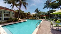 This inviting outdoor amenity space centers around a generously sized freeform swimming pool with crystal-clear turquoise water, designed to evoke a resort-style atmosphere. The pool is framed by a broad, paver-laid sun deck finished in a classic hexagonal pattern, providing ample space for lounging in the abundant Florida sunshine. Bright lime-green chaise lounges and umbrella-shaded seating areas are thoughtfully arranged throughout the deck, offering both sun and shade options for guests.

A striking red pergola structure anchors one end of the pool area, adding an architectural focal point and a shaded gathering spot. Tall, swaying palm trees and lush tropical greenery surround the space on all sides, creating a secluded, resort-like ambiance while allowing open views of the sky above.

Overlooking the pool is a well-appointed Mediterranean-style clubhouse building, characterized by its terracotta tile roof, sage green exterior, white decorative columns, and large multi-pane windows that allow natural light to fill the interior. The seamless connection between the clubhouse and the pool deck creates a cohesive indoor-outdoor environment. Overall, this space combines tropical landscaping, thoughtful amenities, and warm architectural details to deliver an engaging and relaxing outdoor experience.