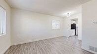 This single-floor space offers a clean, modern aesthetic defined by crisp white walls, light wood-look luxury vinyl plank flooring, and an abundance of natural light throughout. The open living area is generously proportioned, with a large window fitted with blinds that frames outdoor greenery and fills the room with natural light. A flush-mount light fixture adds a subtle decorative touch overhead.

The kitchen is thoughtfully designed with white shaker-style cabinetry, grey quartz countertops, and a full suite of stainless steel appliances, including a refrigerator, electric range, and over-the-range microwave. A door with a window panel at the end of the kitchen provides direct exterior access and additional natural light.

Multiple additional rooms continue the home's consistent neutral palette, each featuring the same light flooring and windows with blinds, creating bright and versatile spaces suitable for a variety of uses. The bathroom is outfitted with classic penny-tile flooring in a cream tone, a white tile tub-surround with a curved shower rod, and oil-rubbed bronze fixtures that lend a timeless character.

Throughout, the space maintains a cohesive, move-in-ready feel with a fresh, minimalist design that balances simplicity with functionality.