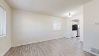 This single-floor space offers a clean, modern aesthetic defined by crisp white walls, light wood-look luxury vinyl plank flooring, and an abundance of natural light throughout. The open living area is generously proportioned, with a large window fitted with blinds that frames outdoor greenery and fills the room with natural light. A flush-mount light fixture adds a subtle decorative touch overhead.

The kitchen is thoughtfully designed with white shaker-style cabinetry, grey quartz countertops, and a full suite of stainless steel appliances, including a refrigerator, electric range, and over-the-range microwave. A door with a window panel at the end of the kitchen provides direct exterior access and additional natural light.

Multiple additional rooms continue the home's consistent neutral palette, each featuring the same light flooring and windows with blinds, creating bright and versatile spaces suitable for a variety of uses. The bathroom is outfitted with classic penny-tile flooring in a cream tone, a white tile tub-surround with a curved shower rod, and oil-rubbed bronze fixtures that lend a timeless character.

Throughout, the space maintains a cohesive, move-in-ready feel with a fresh, minimalist design that balances simplicity with functionality.