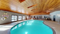 This single-level indoor aquatic facility offers a well-appointed recreational environment designed for relaxation and leisure. The centerpiece is a generously sized, freeform swimming pool with vivid turquoise water, providing ample space for recreational swimming. Adjacent to the main pool sits a separate hot tub, ideal for unwinding after a swim.

The architecture is defined by a striking vaulted wood-planked ceiling with skylights that draw in natural light, creating a warm, lodge-inspired ambiance throughout the space. The walls are clad in textured grey brick, lending a durable yet visually interesting character to the interior. Large windows line the perimeter, allowing abundant natural light to flood the pool deck and offering views of the surrounding exterior.

The pool deck is finished with a durable aggregate flooring and is furnished with multiple lounge chairs and a patio table with seating, providing comfortable spaces to relax poolside. Interior windows along one wall offer a direct view into an adjacent fitness center, seamlessly connecting the two amenity spaces. Wall-mounted sconce lighting adds warmth during evening hours. The facility includes entry and exit doors for convenient access, and signage throughout ensures clear communication of pool rules and safety guidelines.