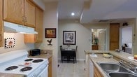 This single-floor residence offers a thoughtfully arranged layout that balances functionality and comfort. The kitchen is a standout feature, equipped with warm honey-toned wood cabinetry that provides ample storage, granite-look countertops, and a white electric range with a ventilation hood overhead. Recessed lighting illuminates the space efficiently, while light tile flooring ties the areas together seamlessly. A structural column elegantly separates the kitchen from the adjacent dining area, creating a natural sense of zone without closing off the open flow between spaces. The dining nook is intimate and inviting, accommodating a small bistro-style table and chairs set beneath framed wall art, making it an ideal spot for casual meals. The neutral beige walls throughout lend a warm, understated ambiance that complements the overall design. A glimpse beyond the dining area reveals additional rooms, including what appears to be a well-lit bathroom with arched architectural detailing and white panel doors, hinting at a refined aesthetic carried throughout the residence. The space is decorated with personal touches, including wall-mounted artwork and decorative accents, giving the home a lived-in, welcoming character.